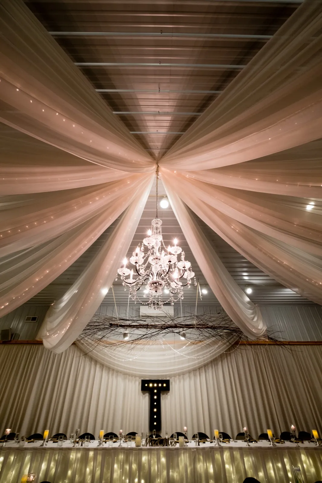 Reception hall ceiling draped with sheer fabric panels converging on a crystal chandelier above a long candlelit head table marked with a lighted marquee T. — Tim Larsen Photography, Brainerd Lakes MN