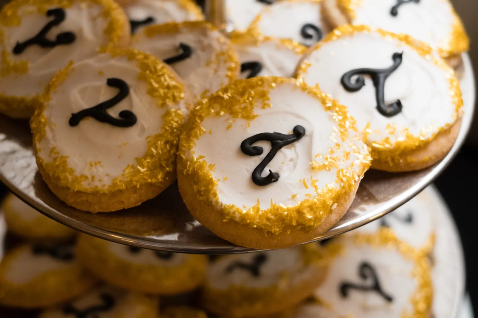 Custom sugar cookies iced in white with the couple's initials piped in black and edges rimmed in gold sanding sugar, arranged on a tiered silver stand. — Tim Larsen Photography, Brainerd Lakes MN