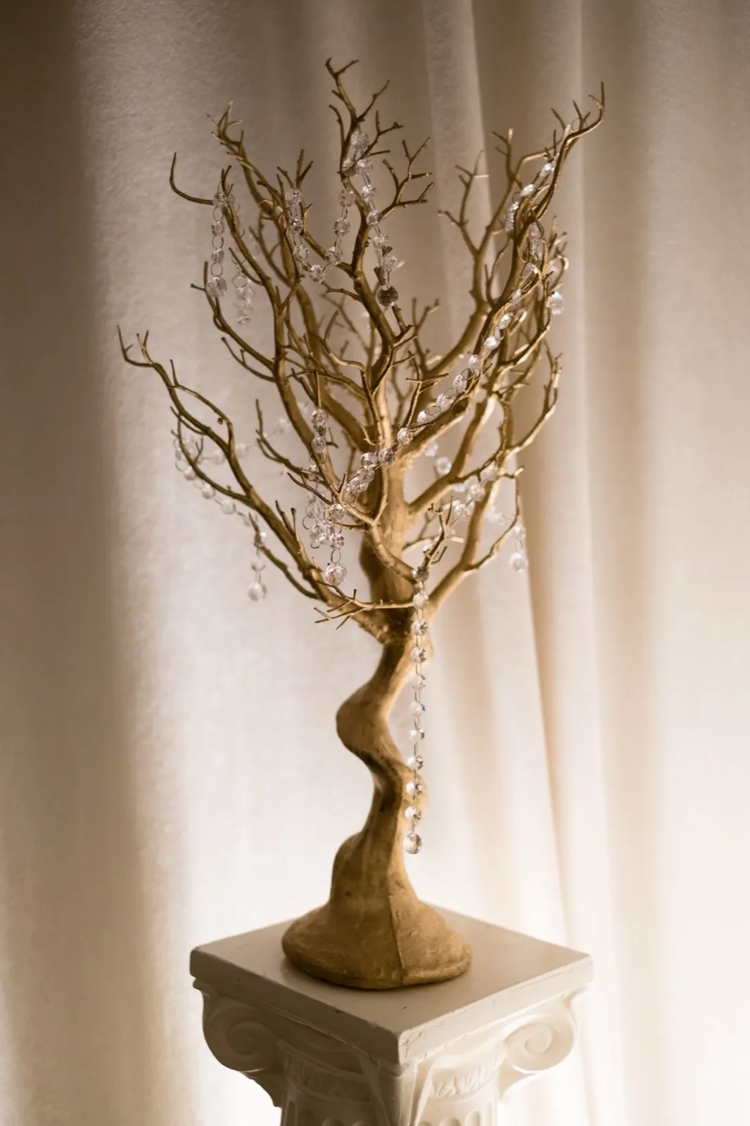 A gilded branch sculpture hung with crystal strands sits on an ornate white pedestal against a soft draped curtain. — Tim Larsen Photography, Brainerd Lakes MN