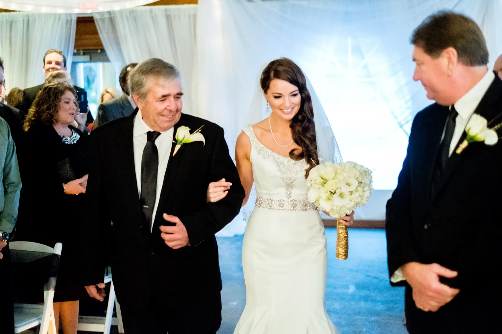 The bride smiles as she walks down the aisle on her father's arm, holding a white bouquet, with a draped fabric ceremony backdrop behind her. — Tim Larsen Photography, Brainerd Lakes MN