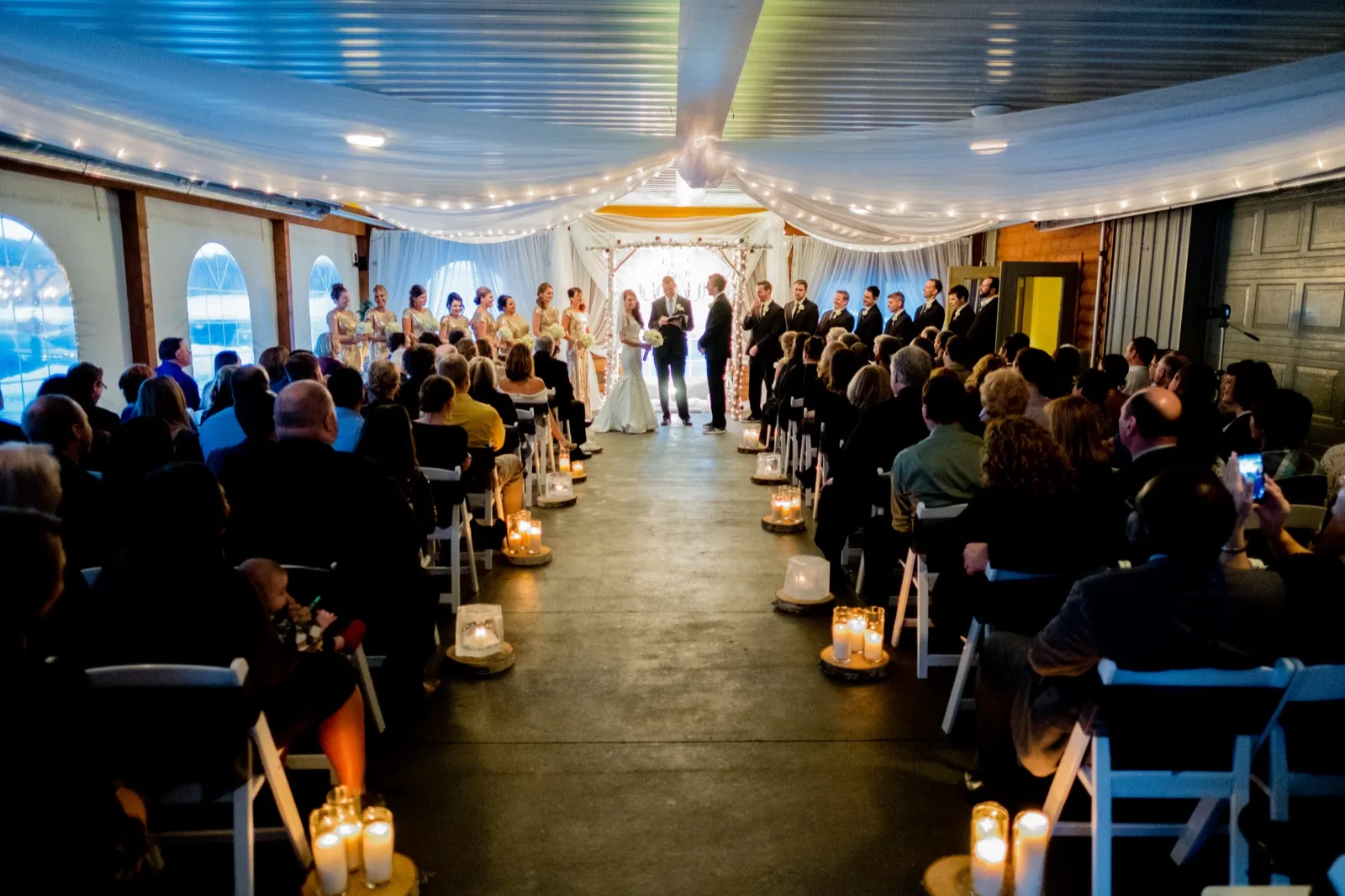 A wide view down the candlelit aisle shows the couple at the altar beneath a draped ceiling and string lights, with the bridal party and seated guests on either side. — Tim Larsen Photography, Brainerd Lakes MN