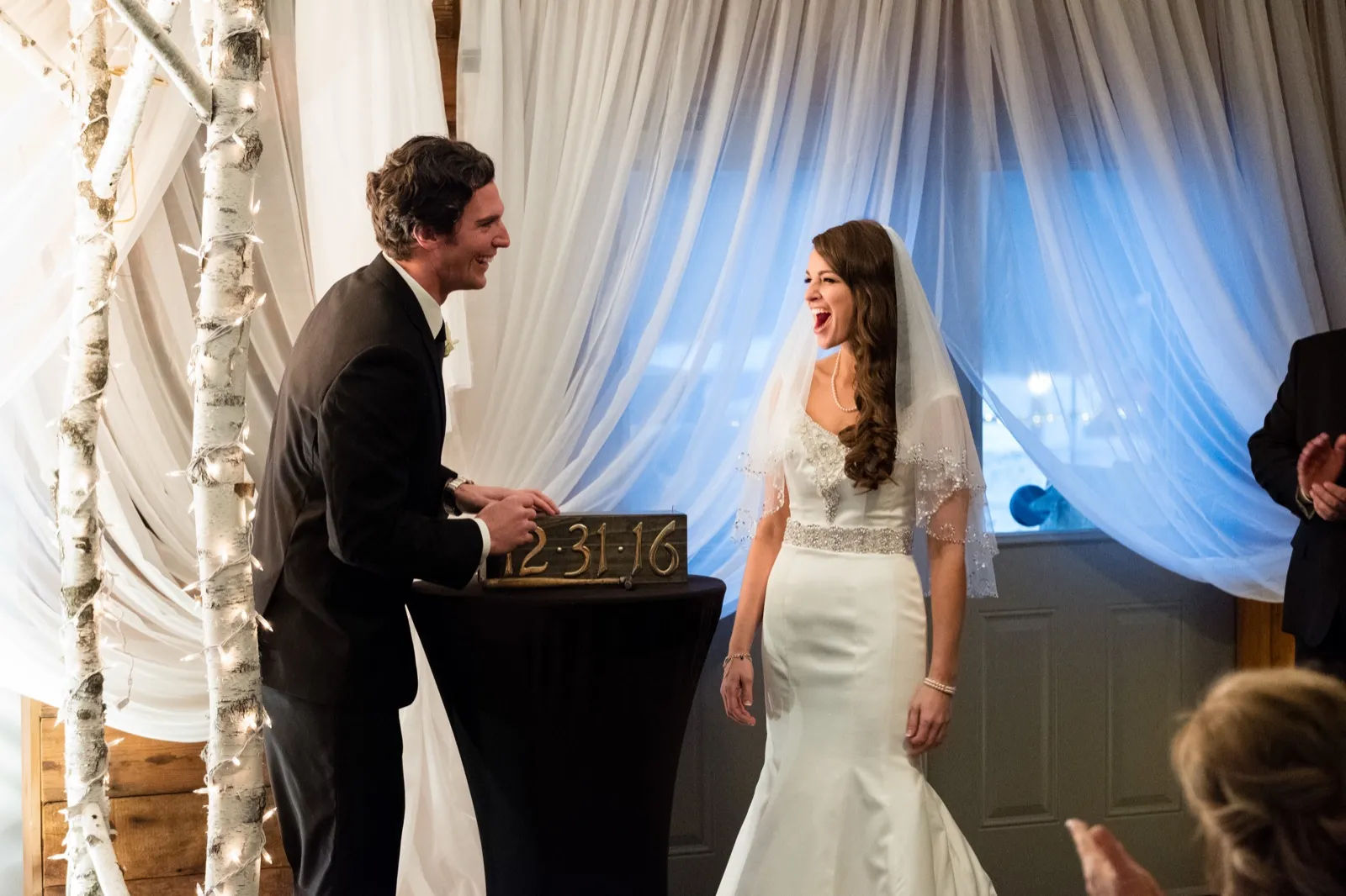 The bride laughs openly during the ceremony as the groom smiles at her across a wooden 12-31-16 date sign at a birch-and-light arch. — Tim Larsen Photography, Brainerd Lakes MN