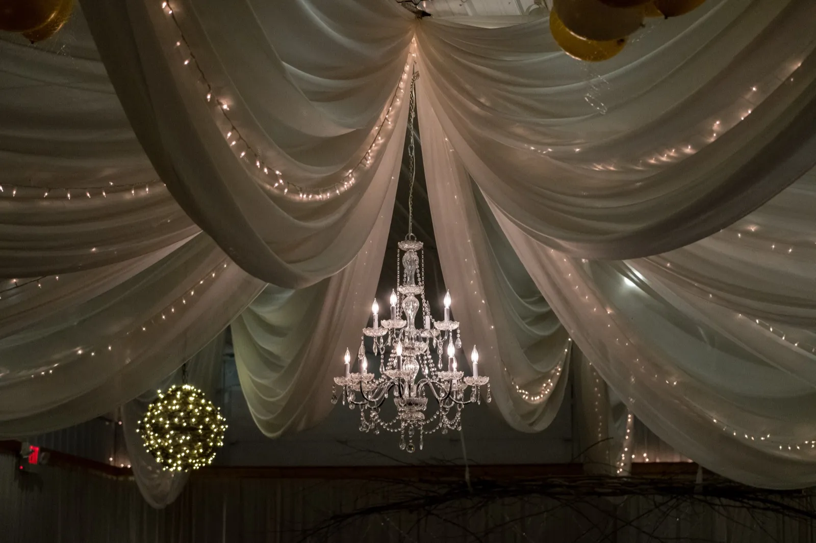 A crystal chandelier hangs from a ceiling draped with white fabric and warm string lights at the Pine Peaks reception space. — Tim Larsen Photography, Brainerd Lakes MN