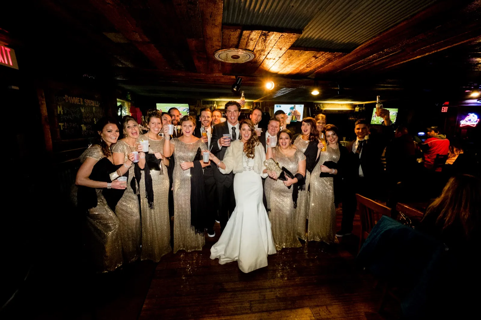The bride and groom stand surrounded by their bridal party in a wood-paneled bar, everyone raising drinks and laughing toward the camera. — Tim Larsen Photography, Brainerd Lakes MN