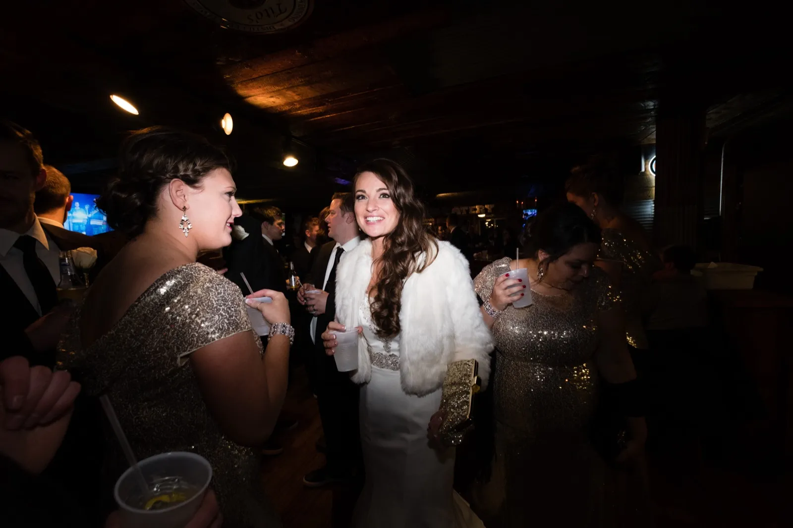 The bride in a white fur stole smiles among her bridesmaids holding drinks in a dim, wood-paneled bar. — Tim Larsen Photography, Brainerd Lakes MN
