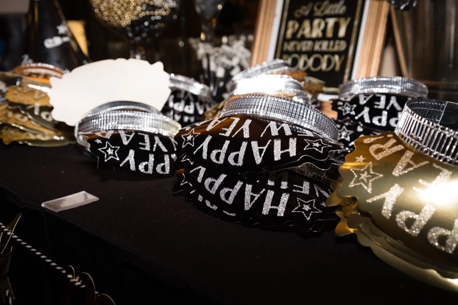 A row of black and gold 'Happy New Year' party hats stacked on a dark reception table beside a framed sign reading 'A Little Party Never Killed Nobody.' — Tim Larsen Photography, Brainerd Lakes MN