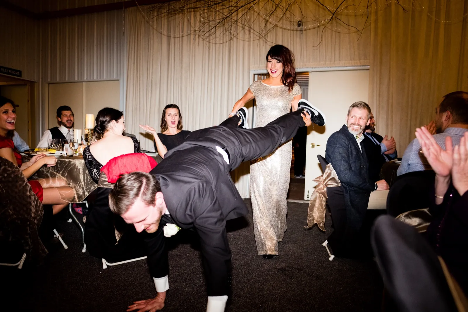 The groom is held horizontal mid-air as the bride in a gold gown holds up his sneakers, guests laughing and clapping around them. — Tim Larsen Photography, Brainerd Lakes MN