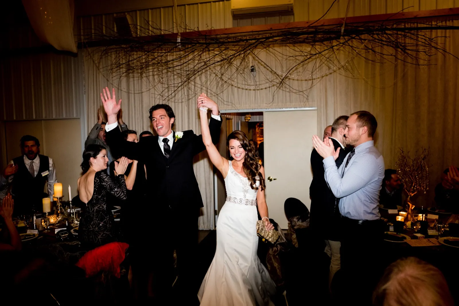 The bride and groom raise their joined hands triumphantly as guests clap on either side during their reception entrance beneath a branch and twinkle-light installation. — Tim Larsen Photography, Brainerd Lakes MN