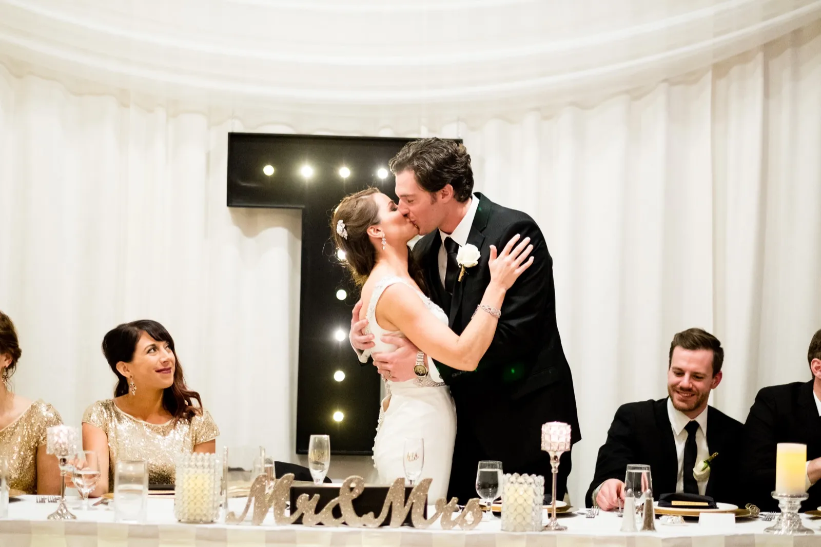 The bride and groom kiss behind a 'Mr & Mrs' sign at the head table while a bridesmaid looks up at them and a groomsman smiles to the side. — Tim Larsen Photography, Brainerd Lakes MN