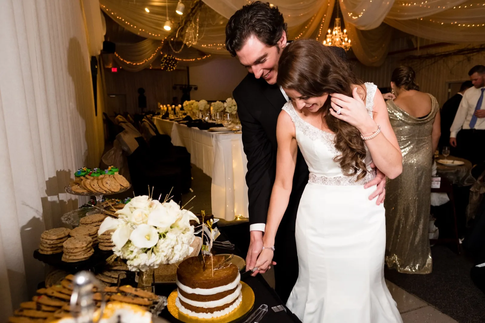 The bride and groom laugh against each other as they cut a small naked layer cake at a reception table lined with cookies. — Tim Larsen Photography, Brainerd Lakes MN