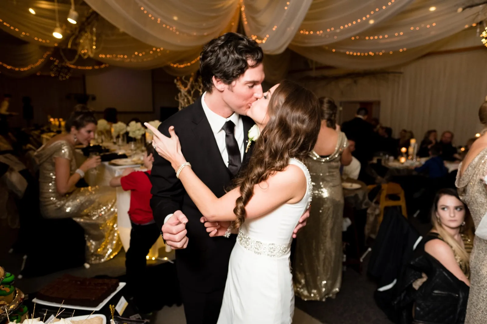 The bride and groom kiss in the middle of the reception room as guests sit at surrounding tables beneath draped fabric and string lights. — Tim Larsen Photography, Brainerd Lakes MN