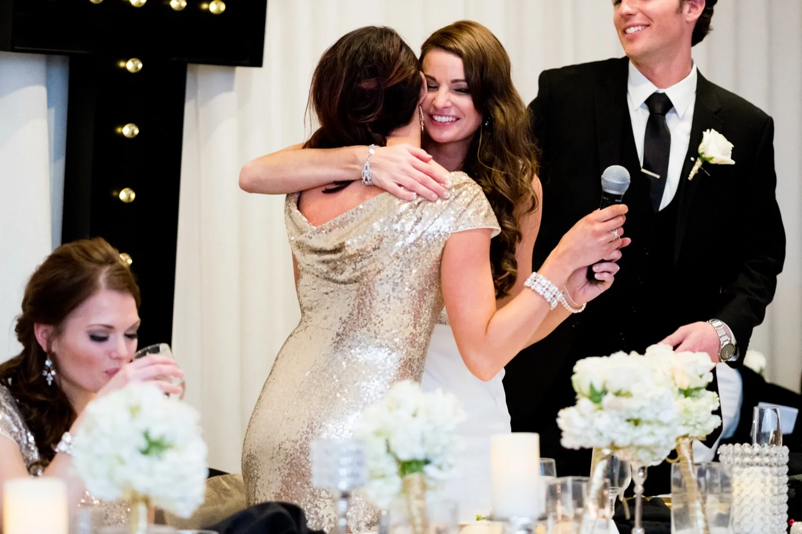 The bride embraces a bridesmaid holding a microphone after a toast while the groom smiles beside them at the head table. — Tim Larsen Photography, Brainerd Lakes MN