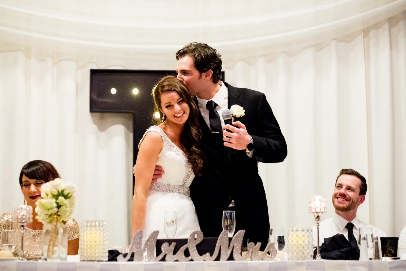The groom kisses the smiling bride on the forehead while holding a microphone during toasts at the head table, with a guest laughing at right. — Tim Larsen Photography, Brainerd Lakes MN