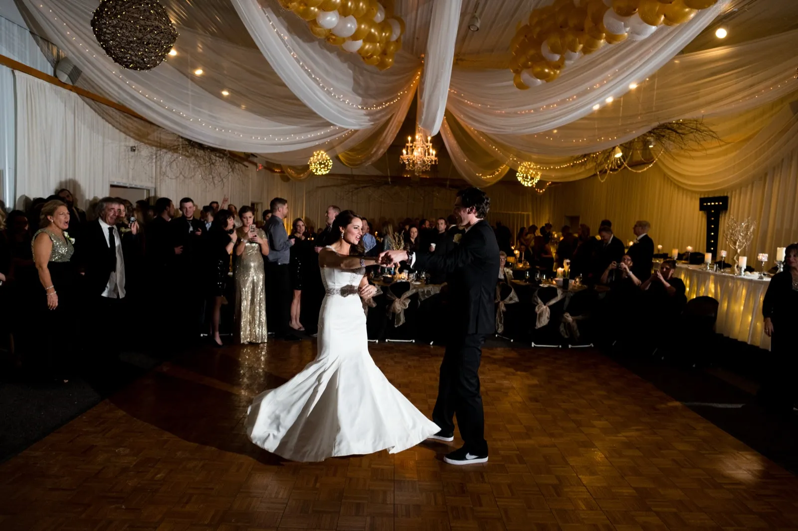 The bride spins on a parquet dance floor as the groom holds her hand, surrounded by guests beneath draped fabric and chandeliers at Pine Peaks Event Center. — Tim Larsen Photography, Brainerd Lakes MN