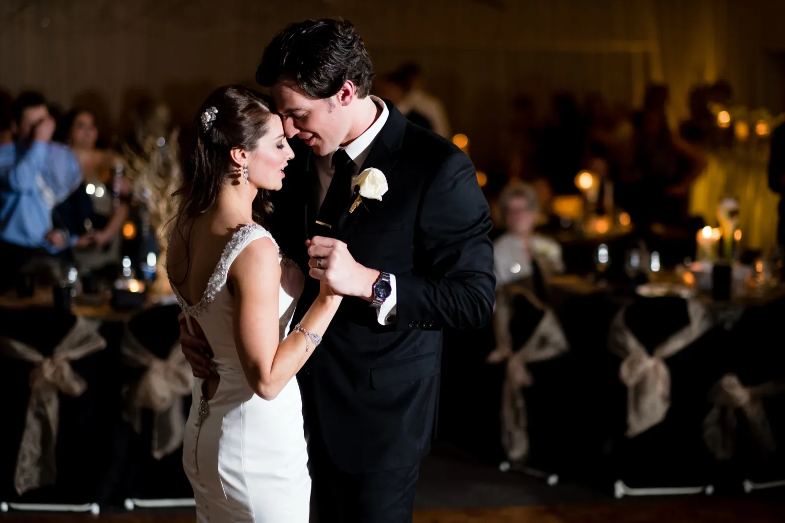 The bride and groom hold each other forehead-to-forehead during their first dance, spotlit on the dance floor with candlelit guest tables glowing behind them. — Tim Larsen Photography, Brainerd Lakes MN
