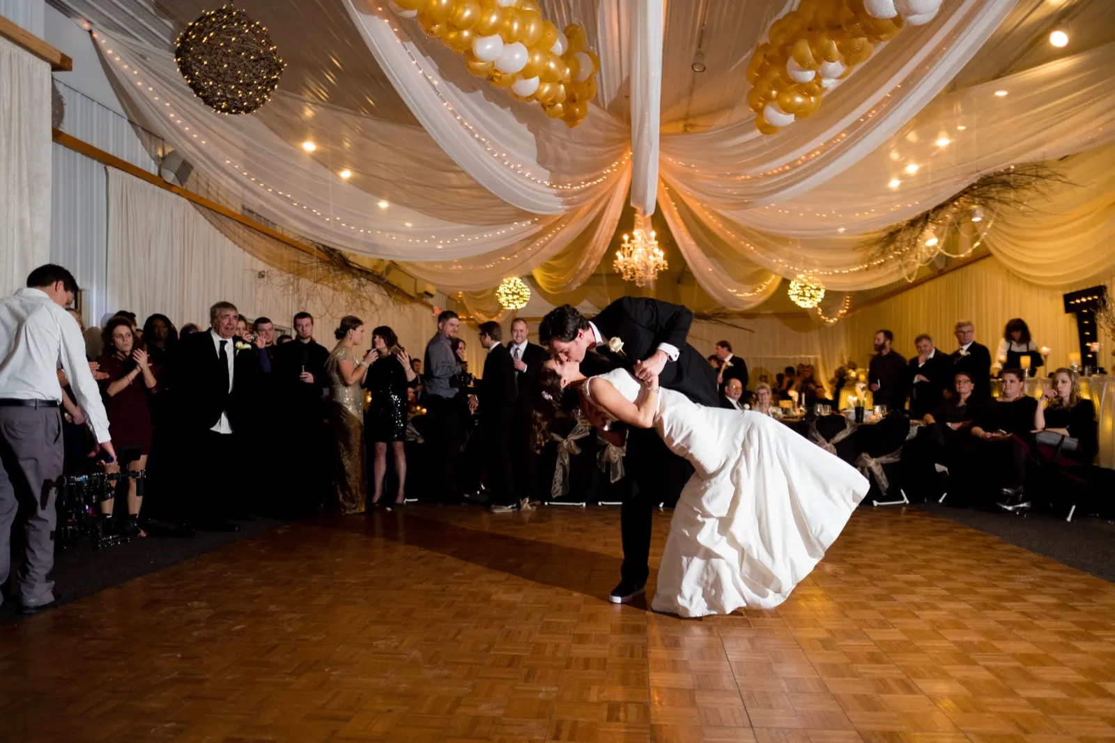 The groom dips the bride into a kiss on the dance floor as guests watch from the edges beneath a draped ceiling and chandelier. — Tim Larsen Photography, Brainerd Lakes MN