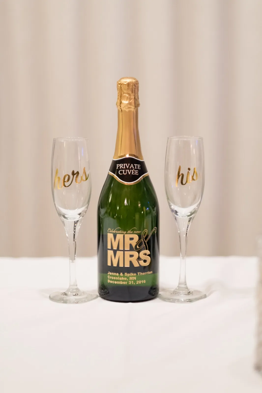A custom-labeled champagne bottle reading 'Mr & Mrs, Jenna & Spike Therrien, Crosslake, MN, December 31, 2016' stands between two gold-script 'his' and 'hers' flutes on a white-linen table. — Tim Larsen Photography, Brainerd Lakes MN