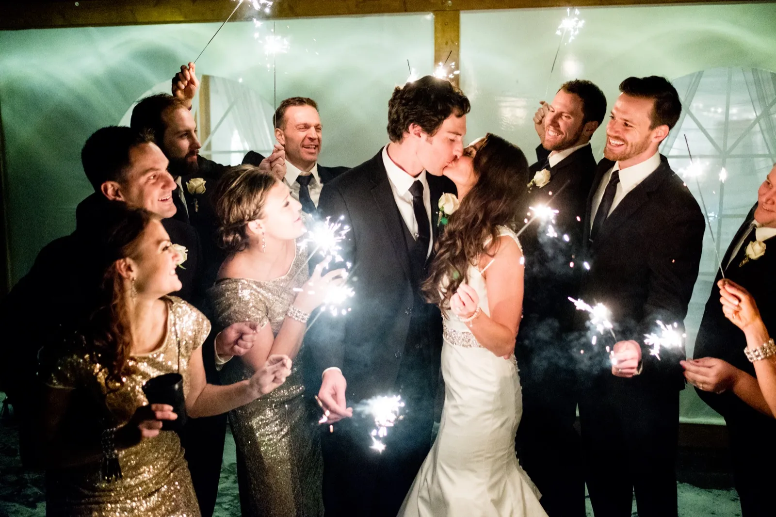 The bride and groom kiss at the center of a circle of wedding party members holding lit sparklers under a covered pavilion at night. — Tim Larsen Photography, Brainerd Lakes MN