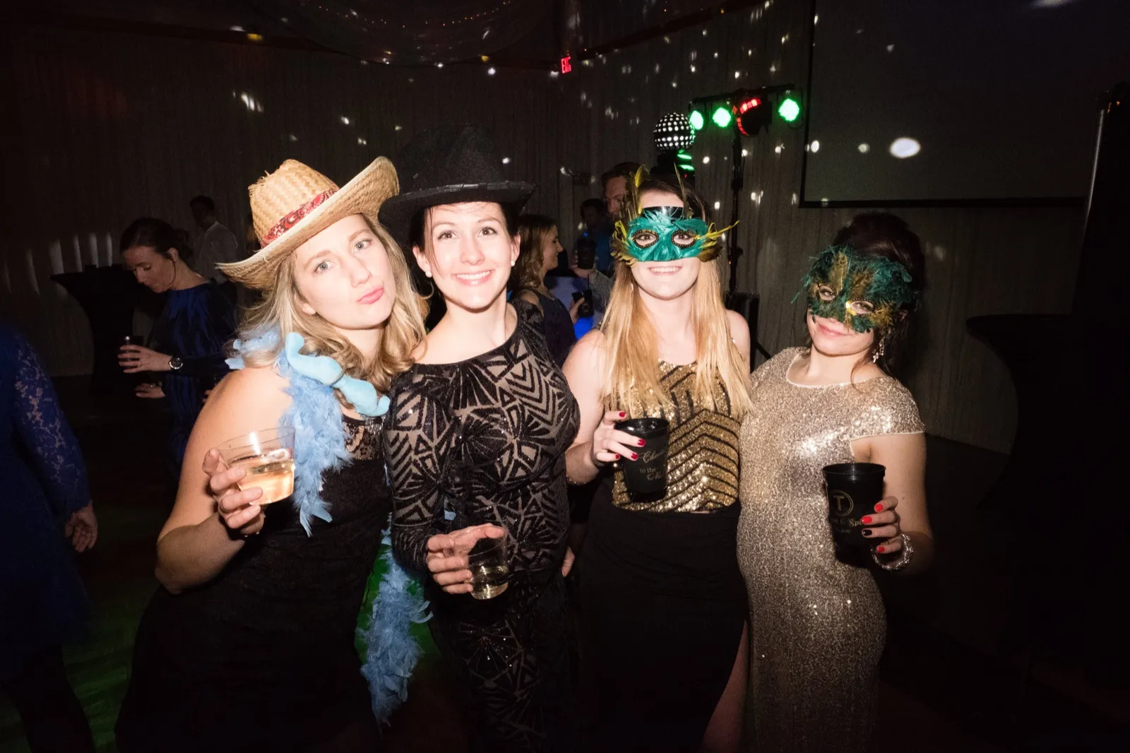 Four wedding guests in cowboy hats, feather boas, and feathered masks pose together on a dimly lit dance floor holding drinks. — Tim Larsen Photography, Brainerd Lakes MN