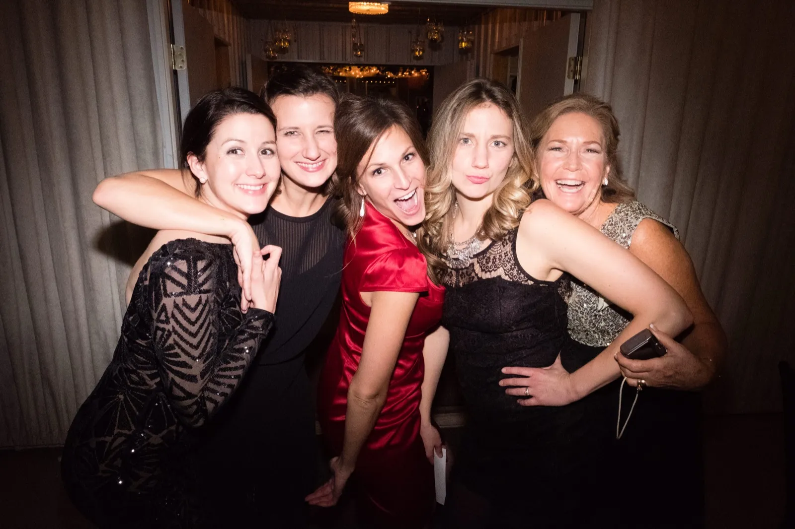 Five women in formal black and red dresses lean into a tight group hug, laughing at the camera in a curtained hallway lit by on-camera flash. — Tim Larsen Photography, Brainerd Lakes MN