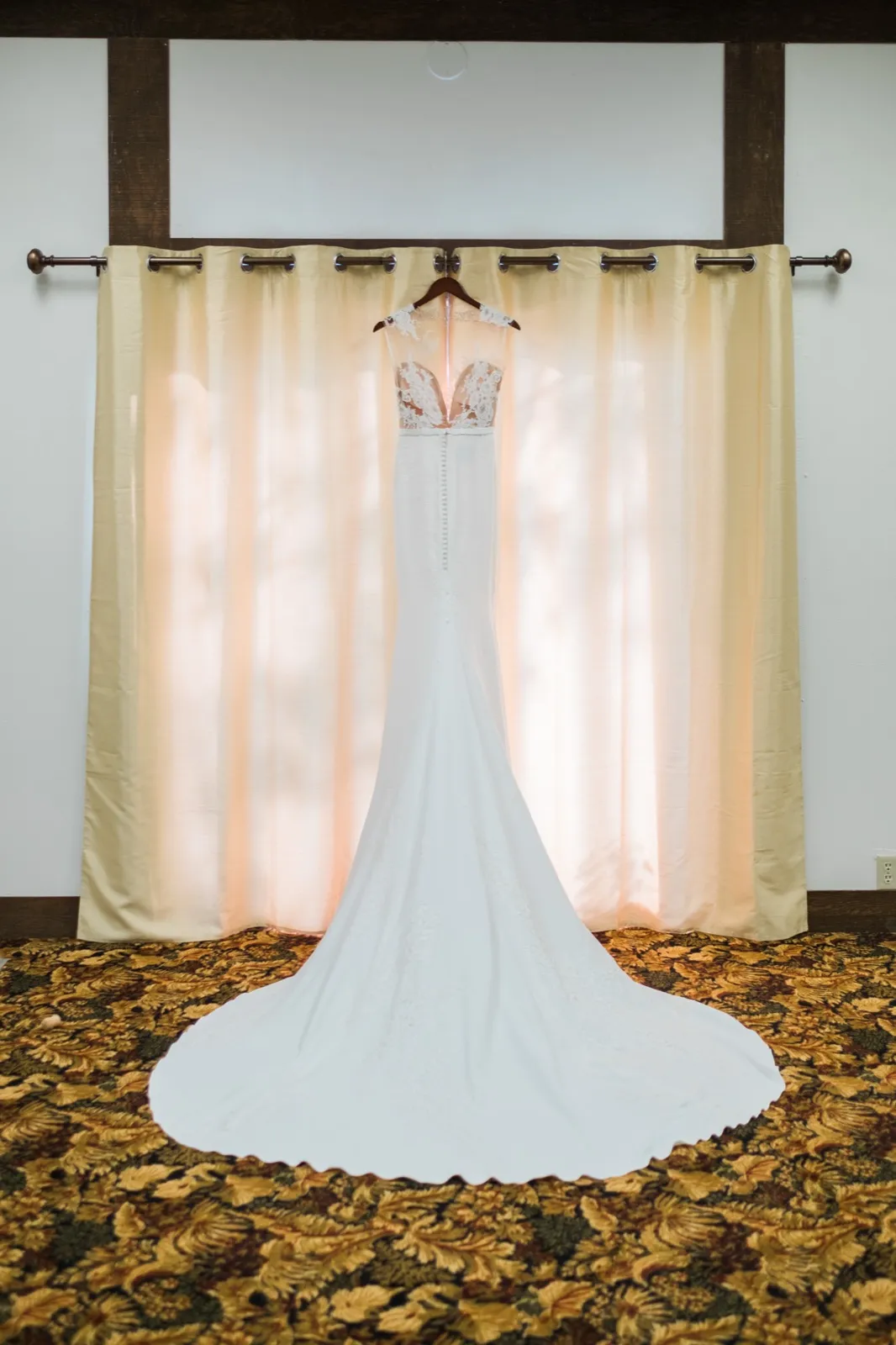 The bride's white lace gown hangs from a curtain rod, train spread across a floral-patterned carpet, backlit by warm window light. — Tim Larsen Photography, Brainerd Lakes MN
