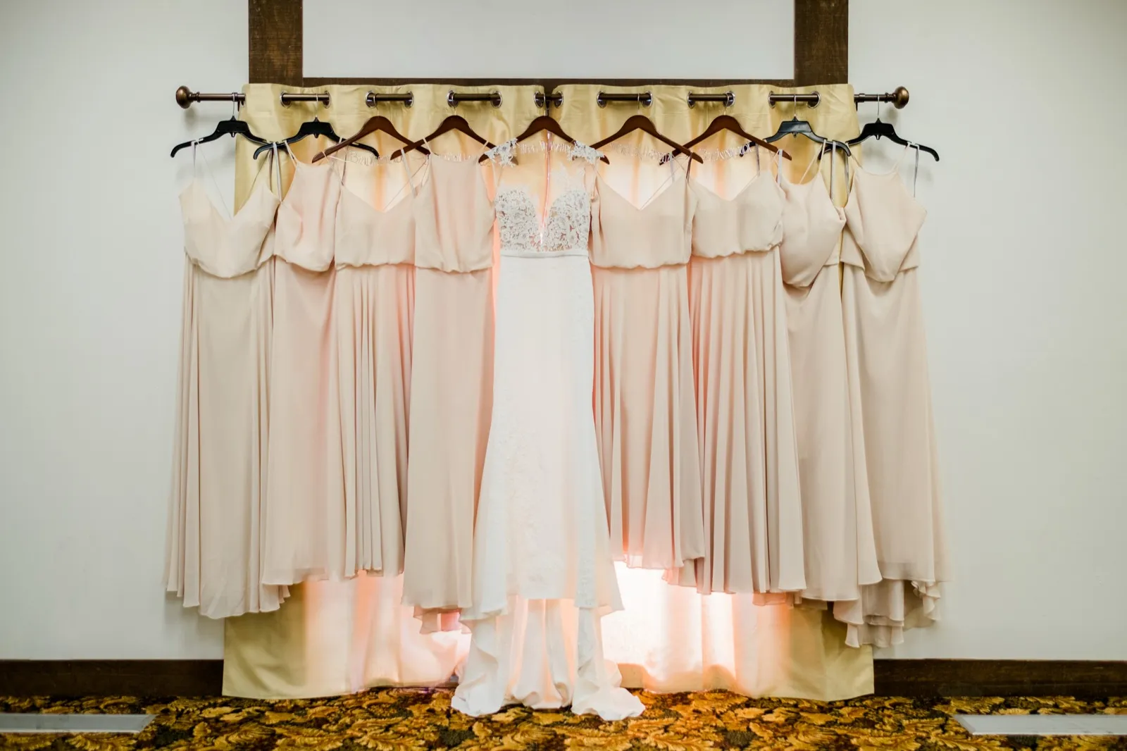 A wedding gown hangs centered between ten blush bridesmaid dresses on a curtain rod, backlit by window light in a bridal suite. — Tim Larsen Photography, Brainerd Lakes MN