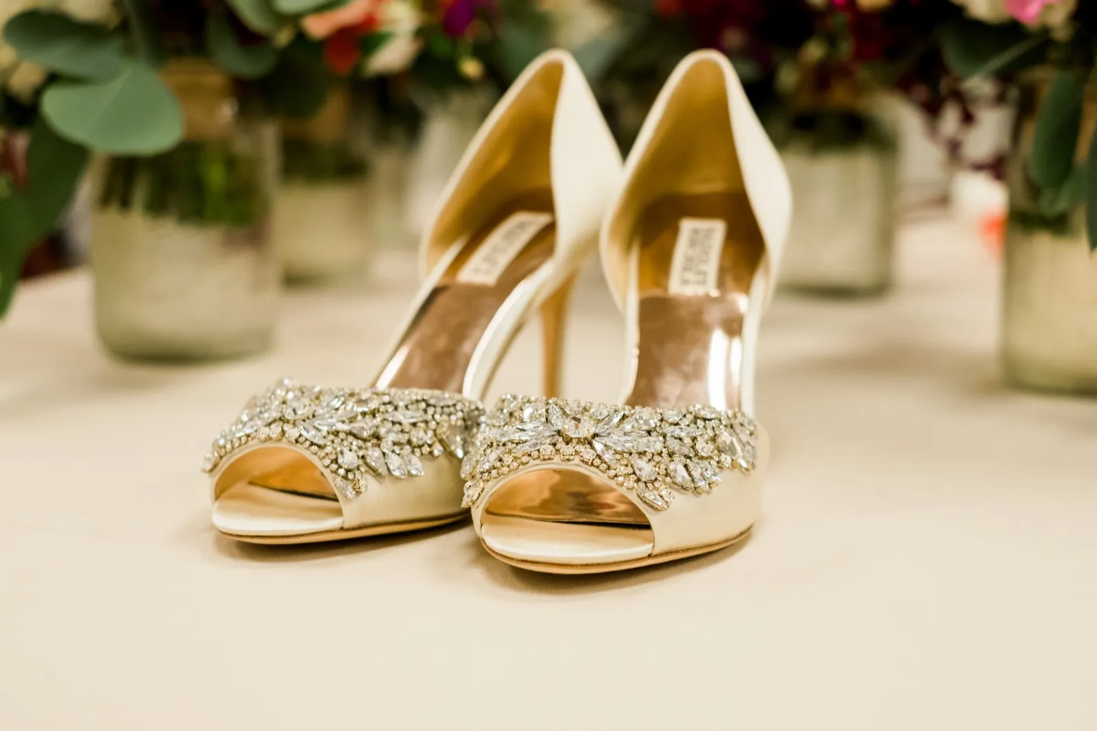 A pair of ivory peep-toe bridal heels with jeweled toe embellishments sits on a table in front of softly blurred floral arrangements. — Tim Larsen Photography, Brainerd Lakes MN