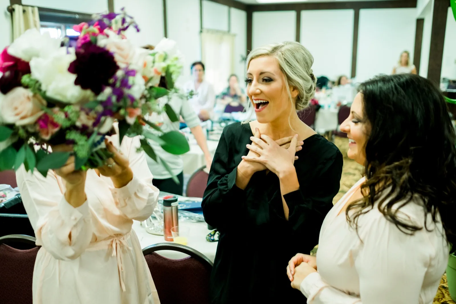 The bride brings both hands to her chest with an open-mouthed reaction as a bridesmaid holds a large bouquet of pink and purple florals into the foreground. — Tim Larsen Photography, Brainerd Lakes MN