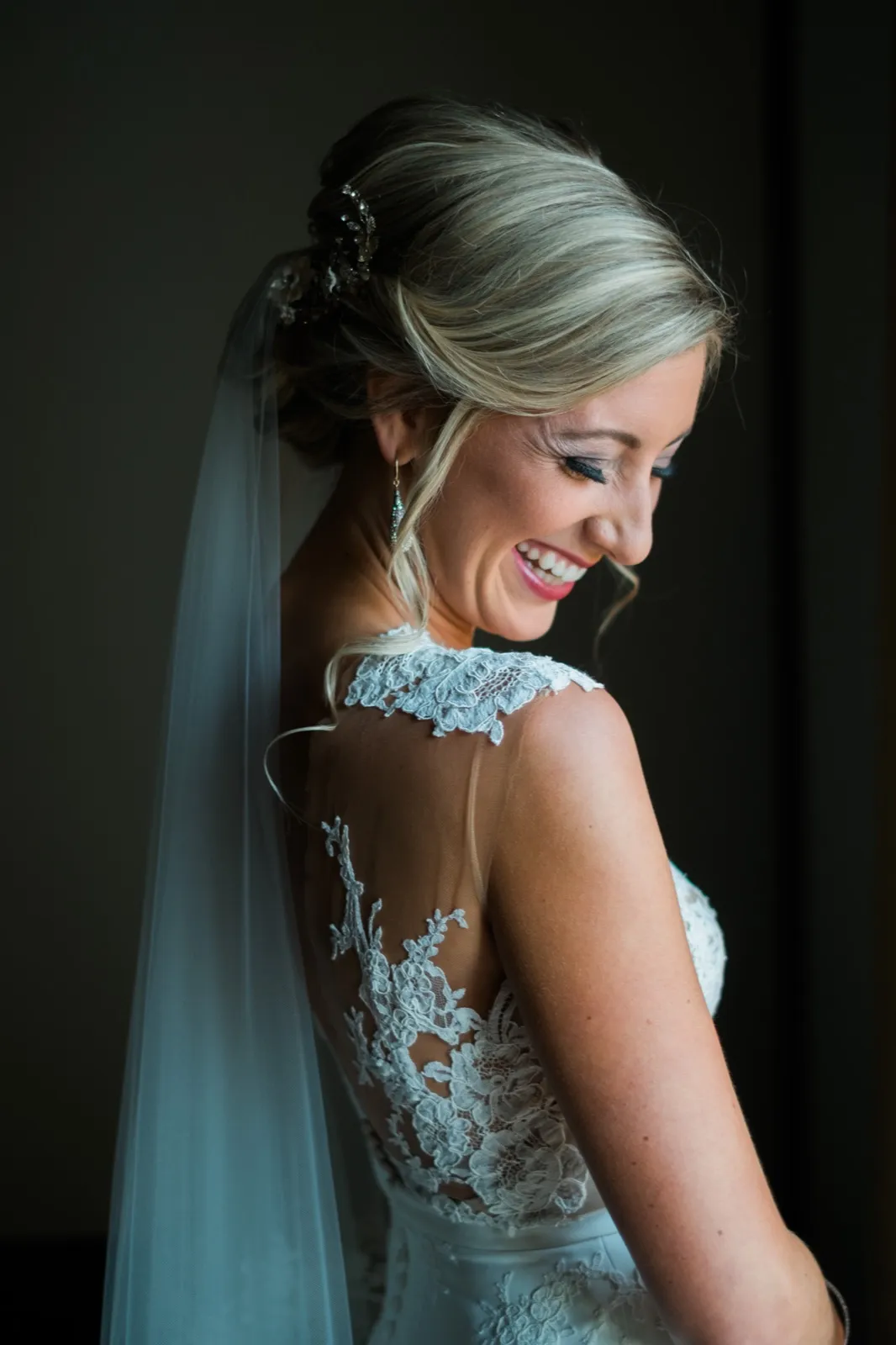 The bride laughs in profile, her veil and lace-back gown illuminated by soft window light against a dark interior. — Tim Larsen Photography, Brainerd Lakes MN