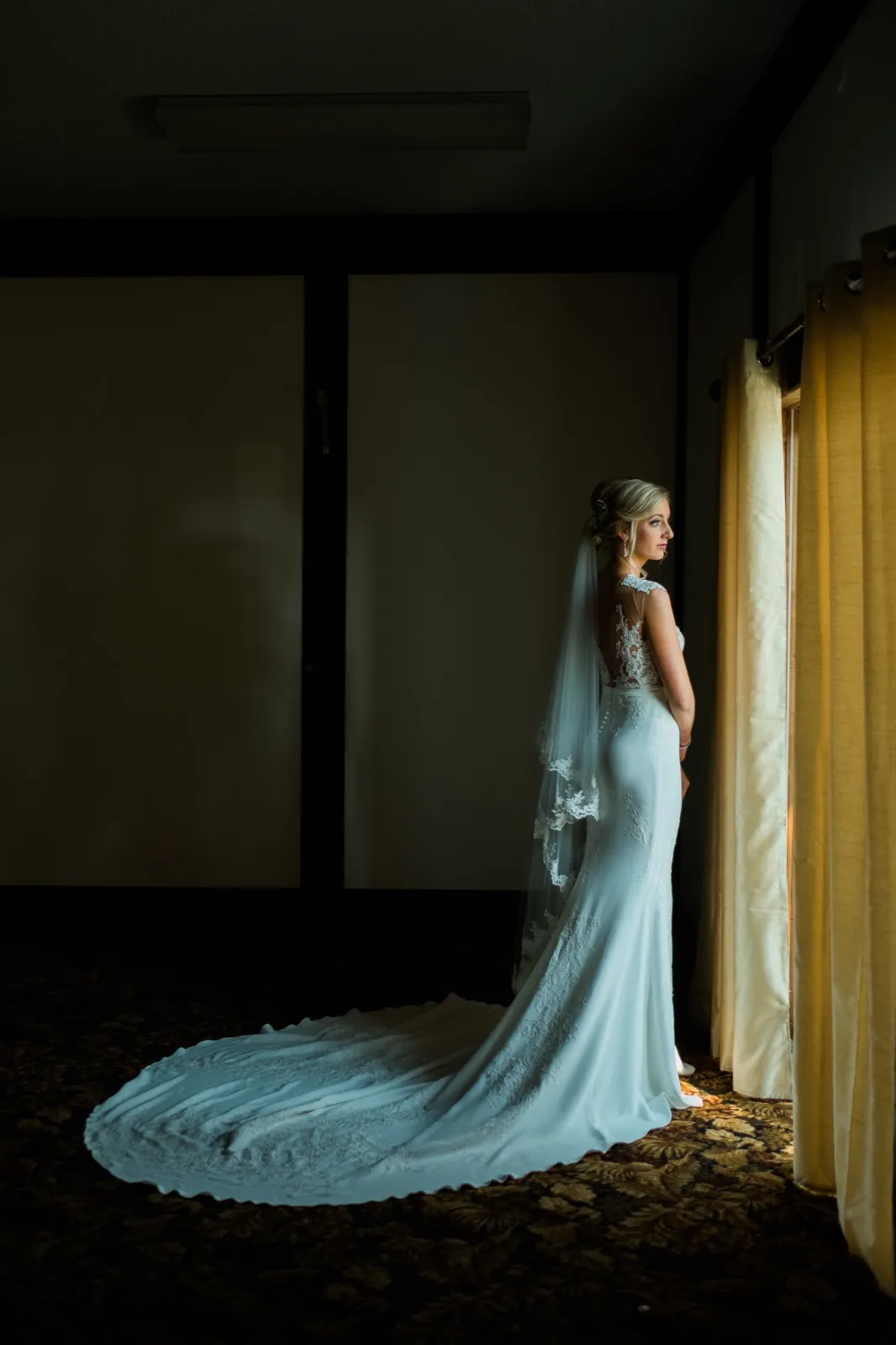 The bride stands in profile beside a tall window, her veil and long train spread across the floor of a darkened bridal suite. — Tim Larsen Photography, Brainerd Lakes MN