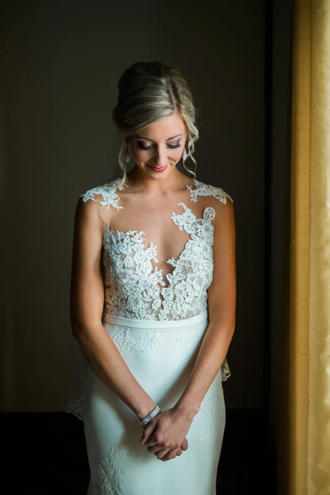 The bride stands in window light against a dark wall, looking down with a soft smile in her lace bodice gown. — Tim Larsen Photography, Brainerd Lakes MN