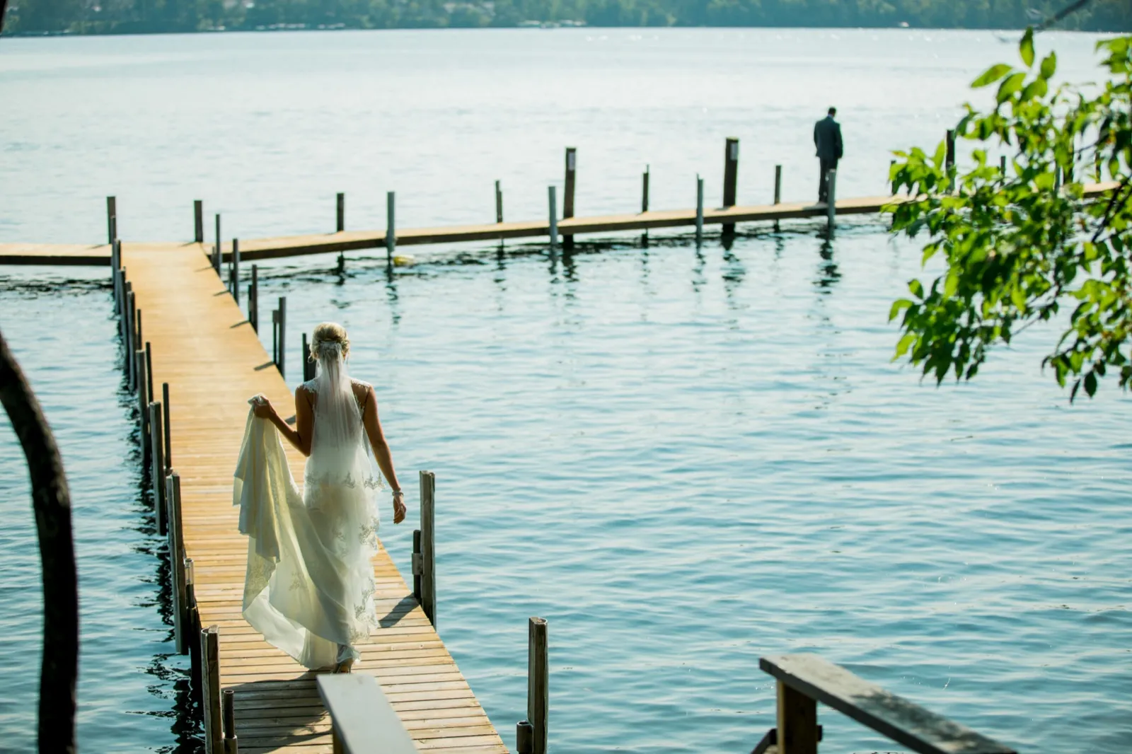 The bride walks down a long wooden dock holding up her gown, approaching the groom who waits with his back turned at the far end over the lake. — Tim Larsen Photography, Brainerd Lakes MN