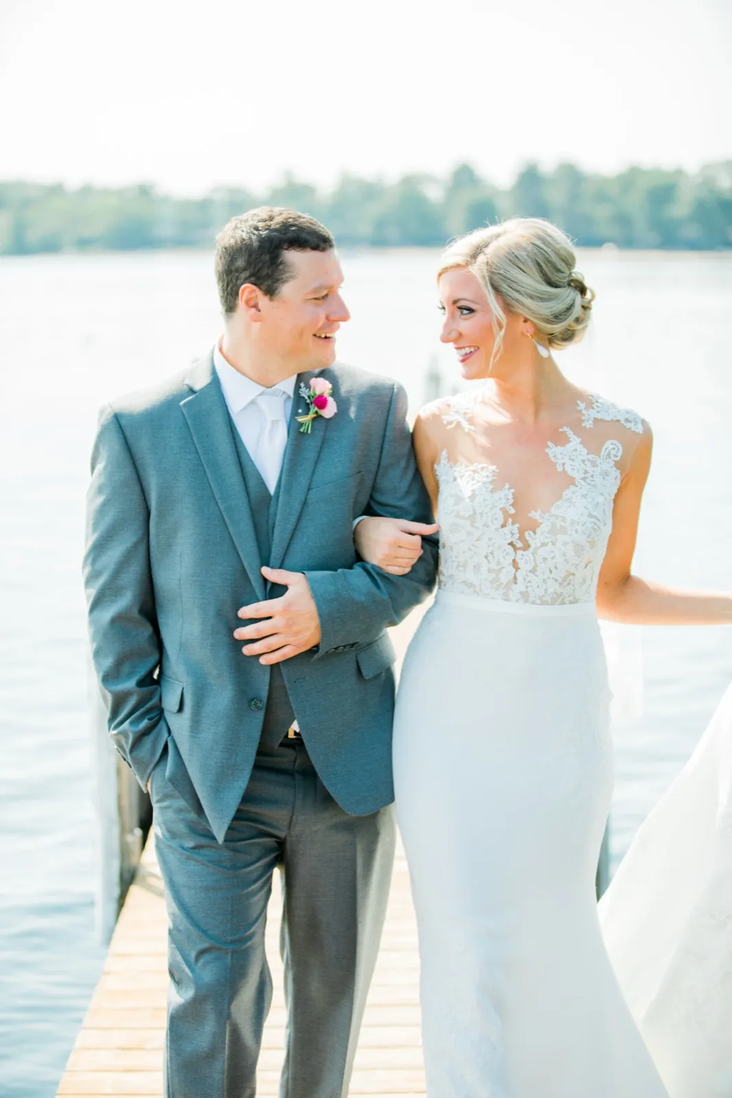 The bride and groom stand arm-in-arm on a wooden dock, laughing as they look at each other with the lake stretching behind them. — Tim Larsen Photography, Brainerd Lakes MN