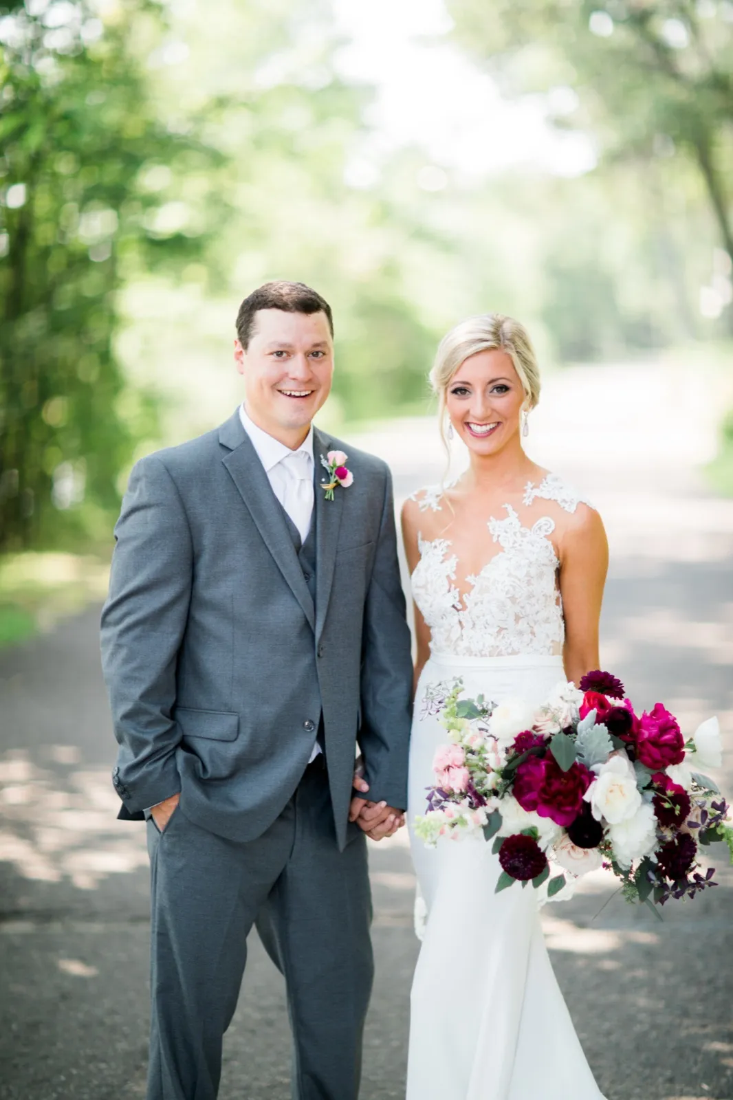 The bride and groom stand holding hands on a tree-lined road, the bride holding a large burgundy and blush bouquet while both smile at the camera. — Tim Larsen Photography, Brainerd Lakes MN