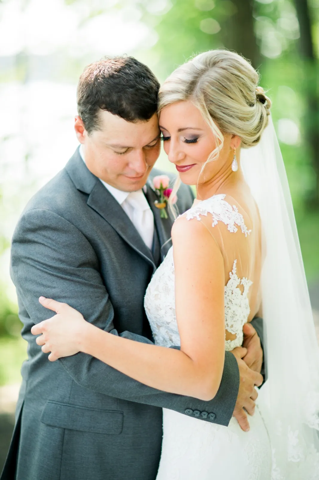 The bride and groom hold each other closely with eyes closed, her veil and lace gown softly lit against blurred green foliage. — Tim Larsen Photography, Brainerd Lakes MN
