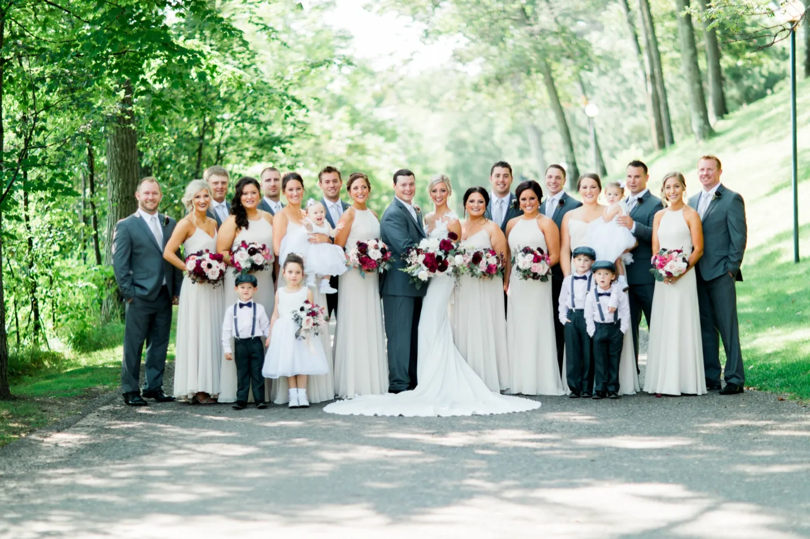 The bride and groom stand at the center of their full bridal party, including flower girls and ring bearers, on a tree-lined path with dappled sunlight overhead. — Tim Larsen Photography, Brainerd Lakes MN