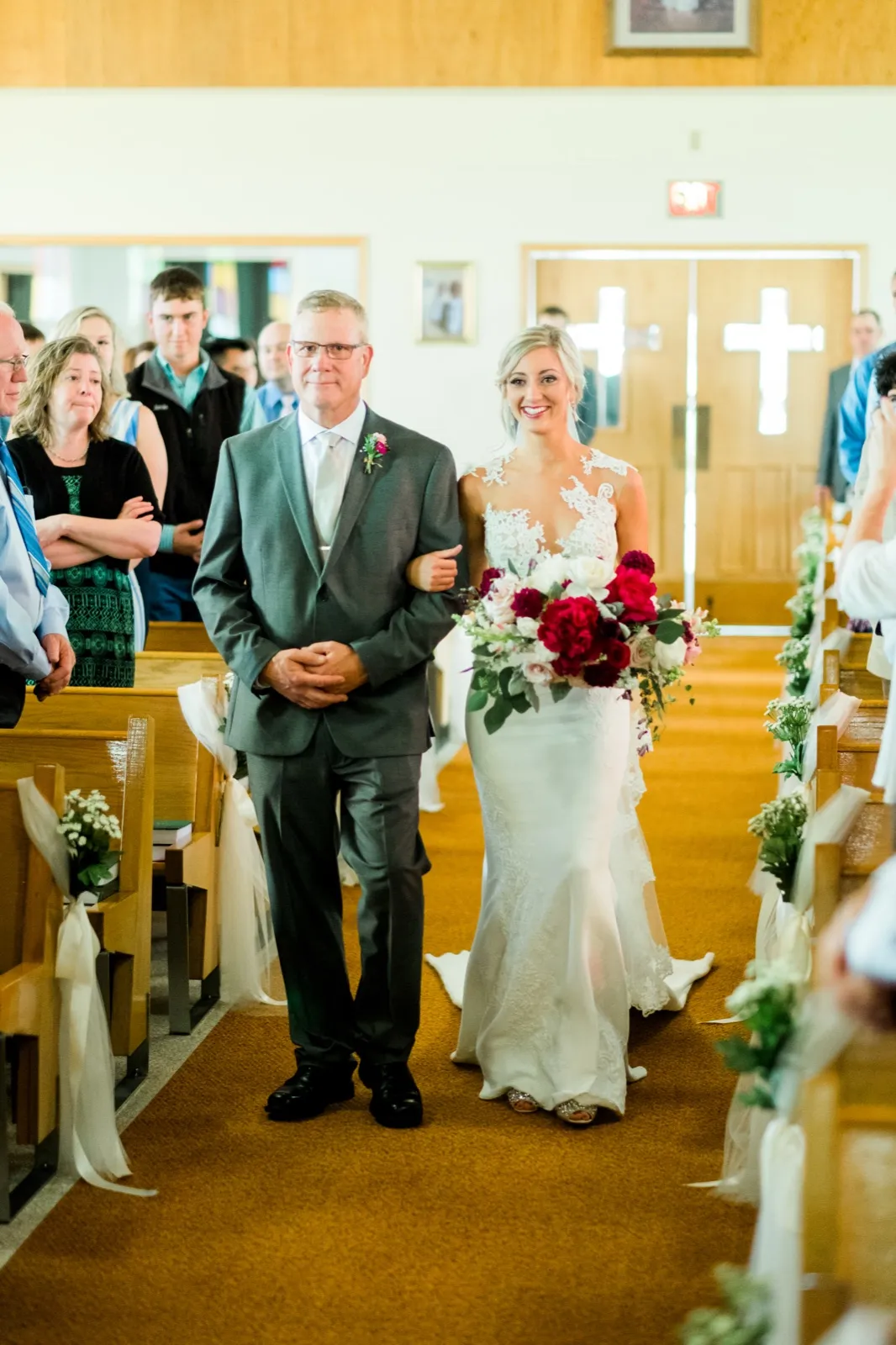 The bride walks down the church aisle on her father's arm, holding a large burgundy and blush bouquet, with guests watching from the pews on either side. — Tim Larsen Photography, Brainerd Lakes MN
