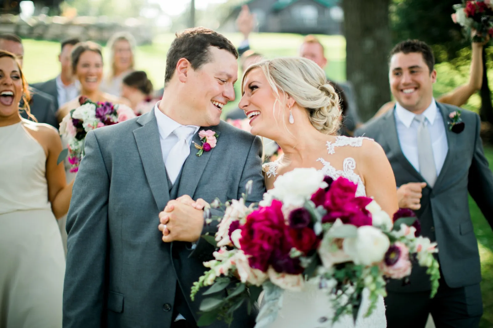 The bride and groom laugh face-to-face as their bridal party cheers around them on a sunlit lawn, jewel-toned bouquets raised in celebration. — Tim Larsen Photography, Brainerd Lakes MN