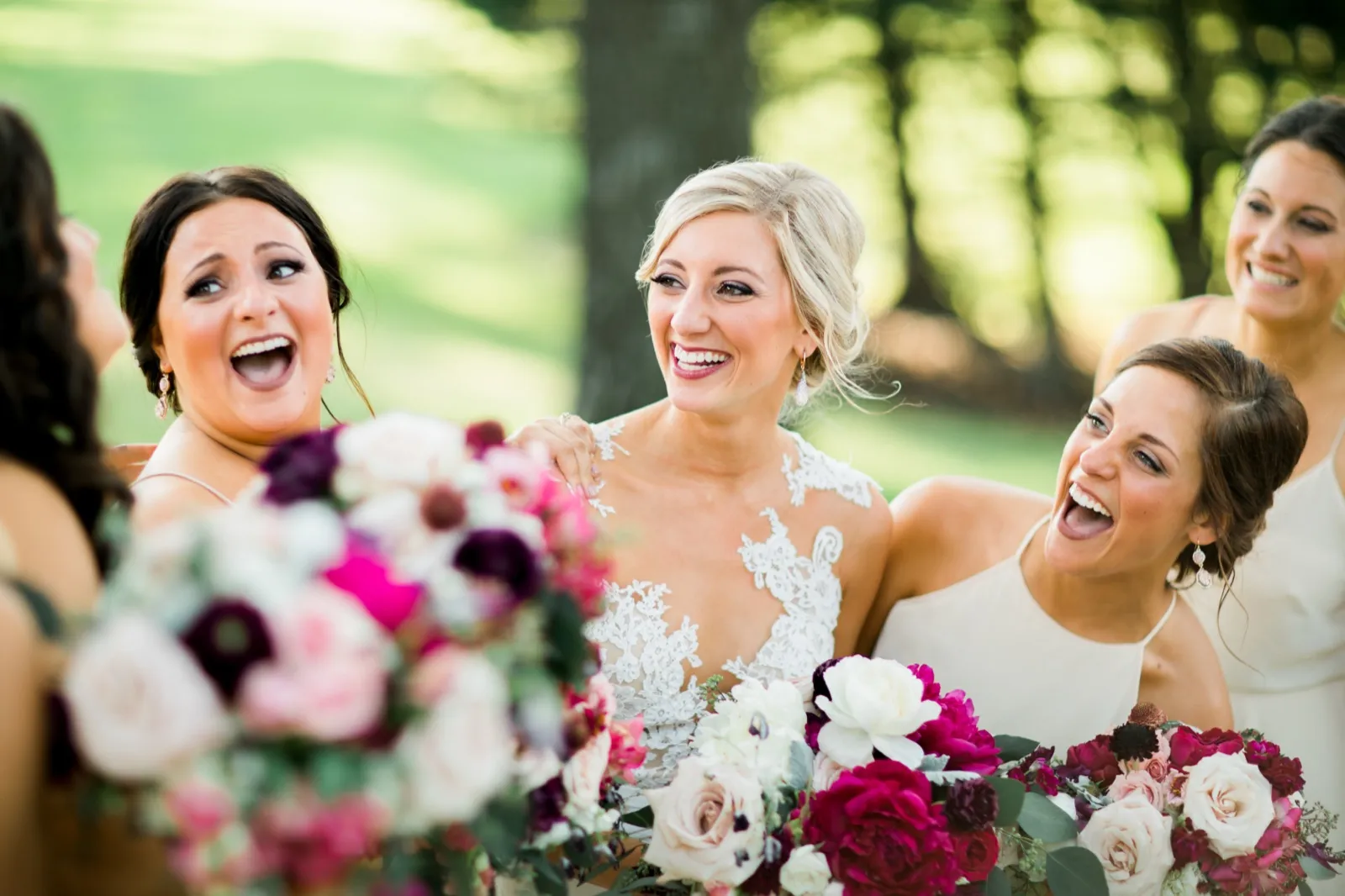The bride laughs with two bridesmaids while a jewel-toned bouquet blurs in the foreground on a tree-shaded lawn. — Tim Larsen Photography, Brainerd Lakes MN