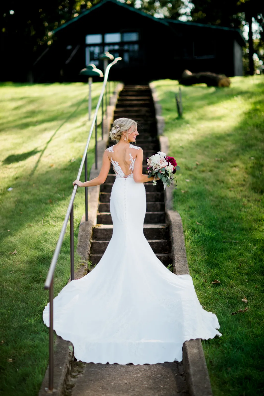 The bride glances over her shoulder while standing on a stone stairway, her gown's train cascading down the steps toward the camera with a lodge building above. — Tim Larsen Photography, Brainerd Lakes MN
