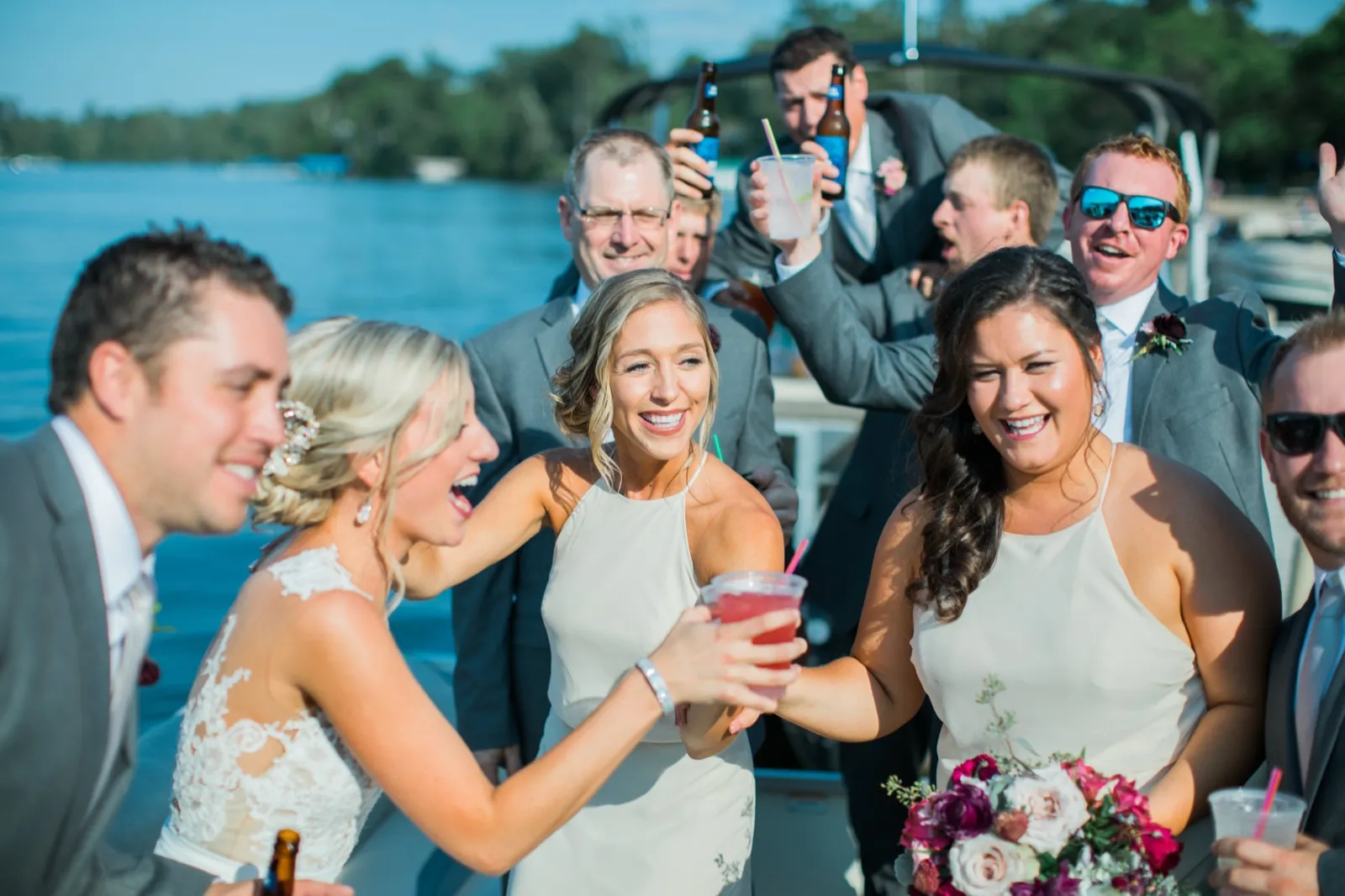 The bride and a bridesmaid clink drinks and laugh on a pontoon boat as groomsmen raise beers behind them on a sunlit lake. — Tim Larsen Photography, Brainerd Lakes MN
