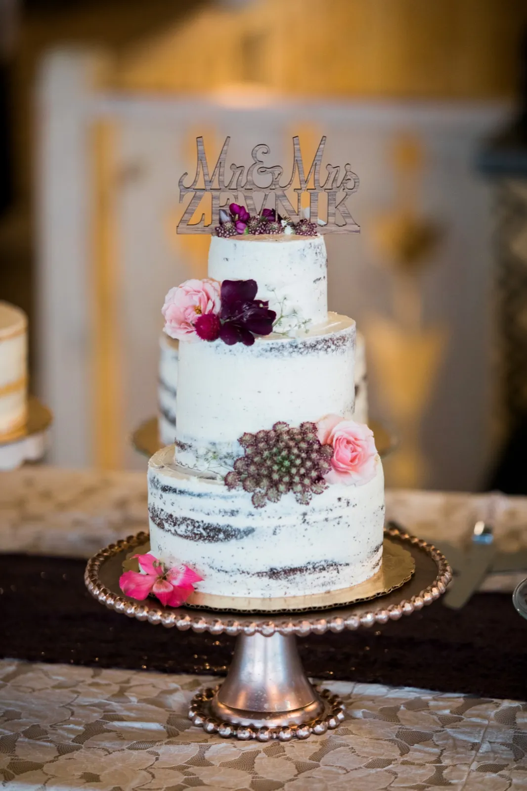 A three-tier naked wedding cake topped with a wooden 'Mr & Mrs Zevnik' sign and decorated with pink and burgundy florals, displayed on a beaded silver pedestal stand. — Tim Larsen Photography, Brainerd Lakes MN