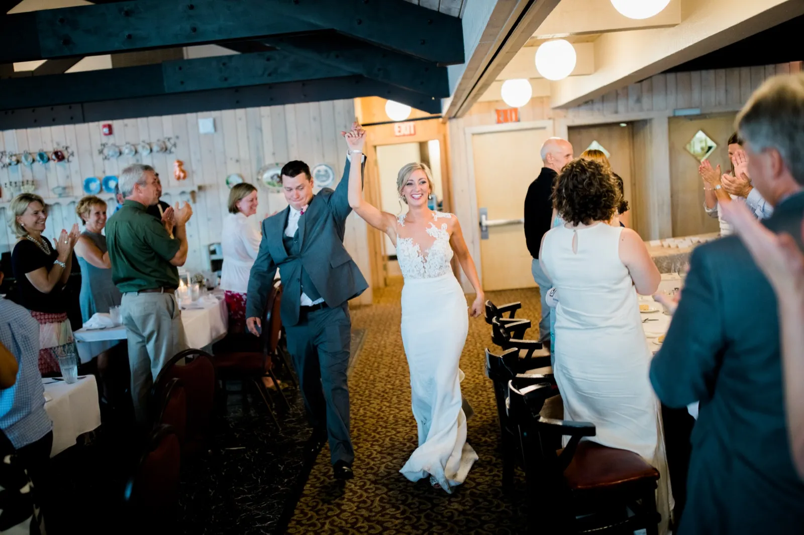 The bride beams as the groom raises her arm triumphantly while walking into the reception space, with seated guests applauding on either side. — Tim Larsen Photography, Brainerd Lakes MN