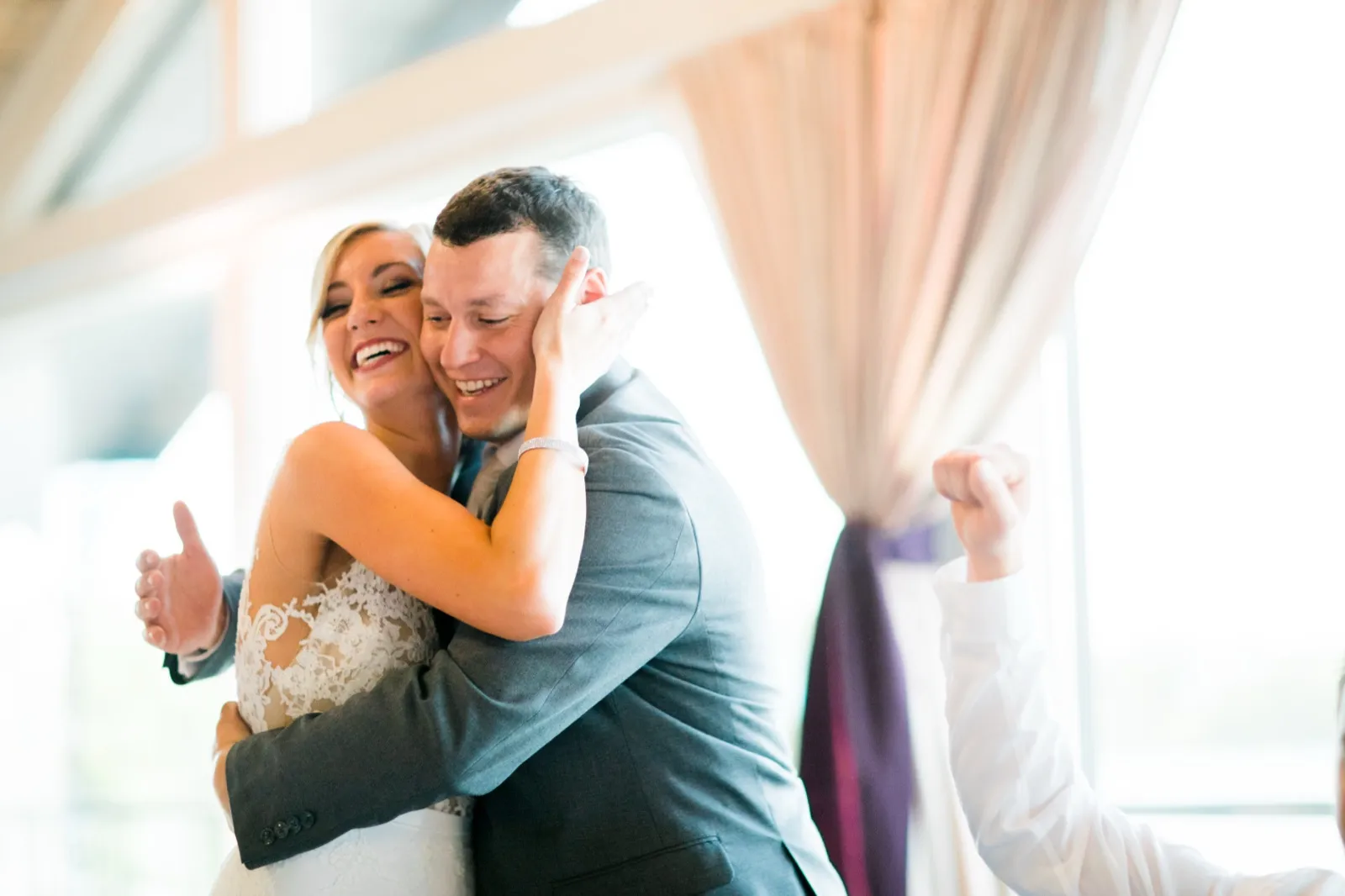 The bride laughs with her eyes closed as the groom pulls her in close on the dance floor, with a cheering guest's raised fist visible at the edge of the frame. — Tim Larsen Photography, Brainerd Lakes MN