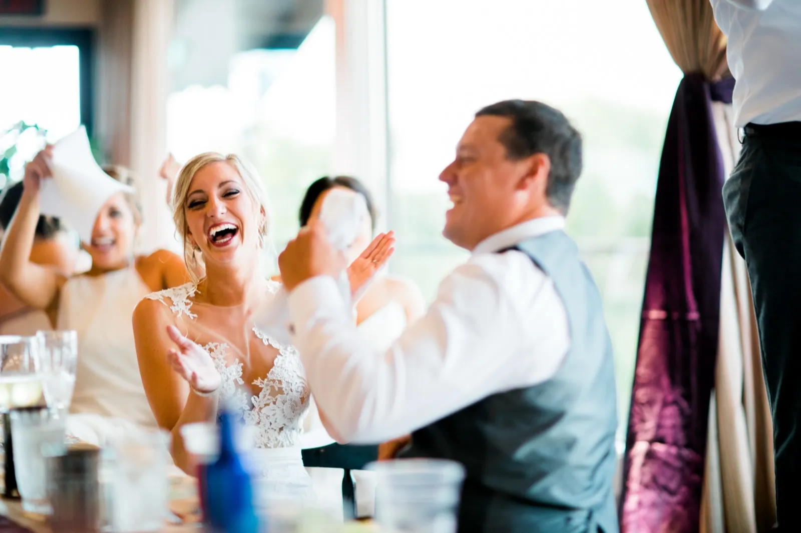 The bride laughs open-mouthed while the groom claps beside her and a bridesmaid waves a napkin overhead during a reception toast. — Tim Larsen Photography, Brainerd Lakes MN