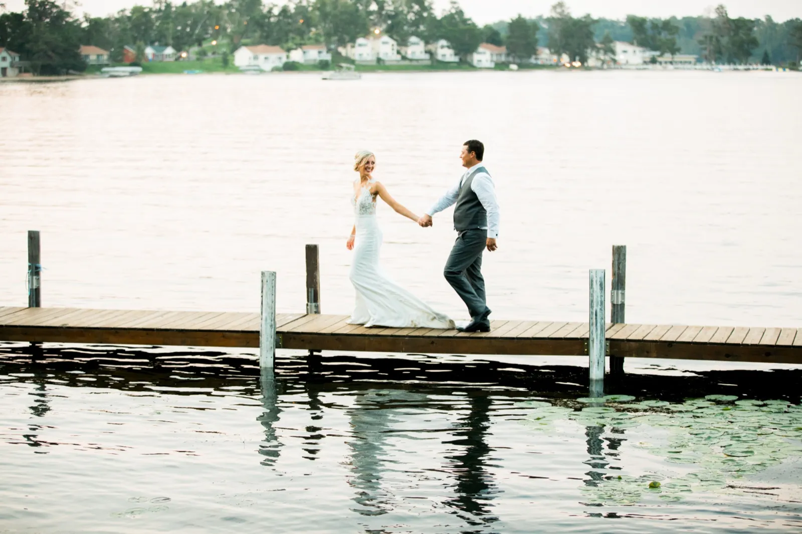 The bride leads the groom by the hand along a long wooden dock over Gull Lake, with the Cragun's Resort shoreline visible in the distance. — Tim Larsen Photography, Brainerd Lakes MN