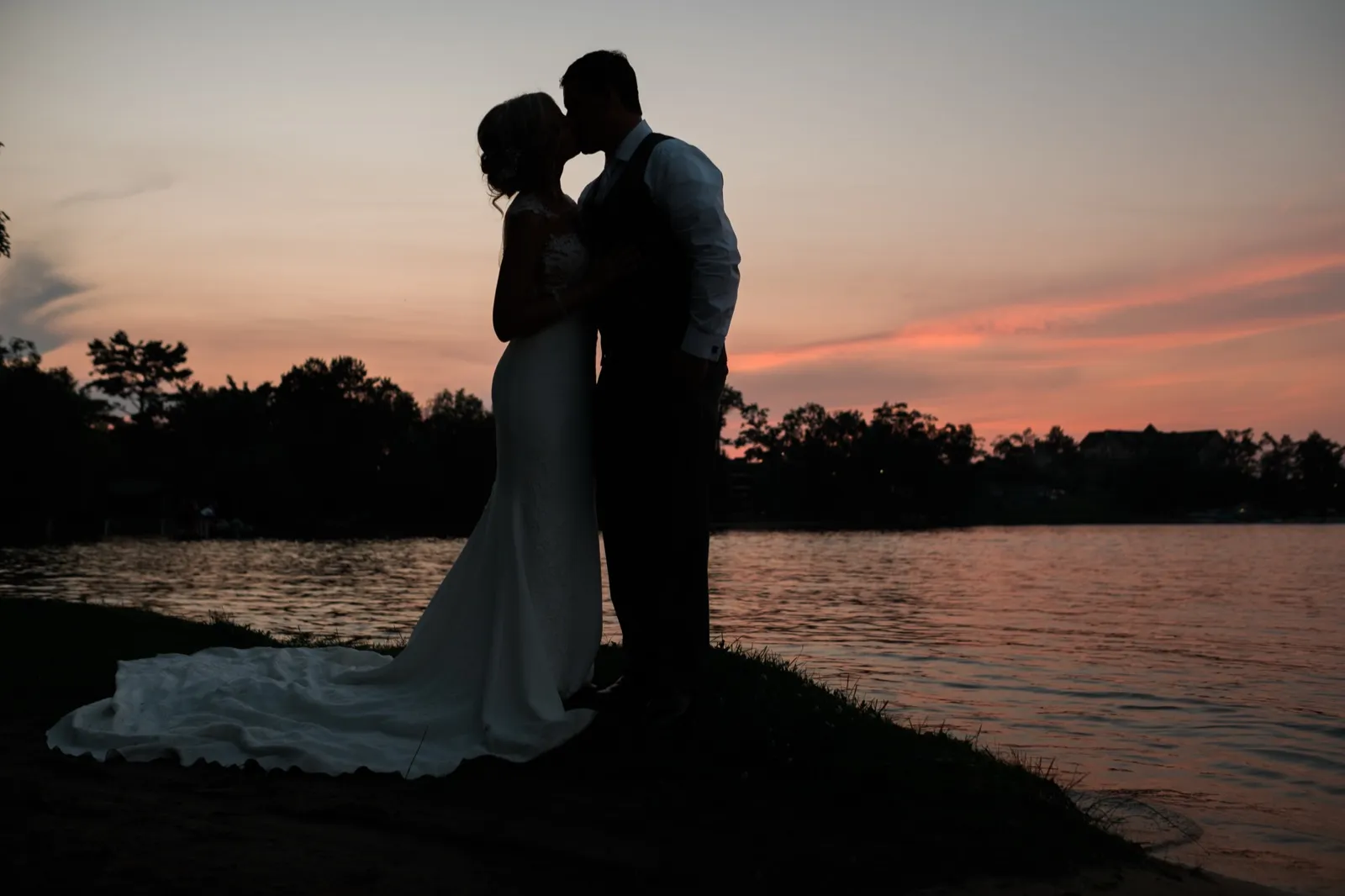 The bride and groom kiss in silhouette on a lakeshore at dusk, her gown's train spilling across the grass against a pink and orange sky. — Tim Larsen Photography, Brainerd Lakes MN