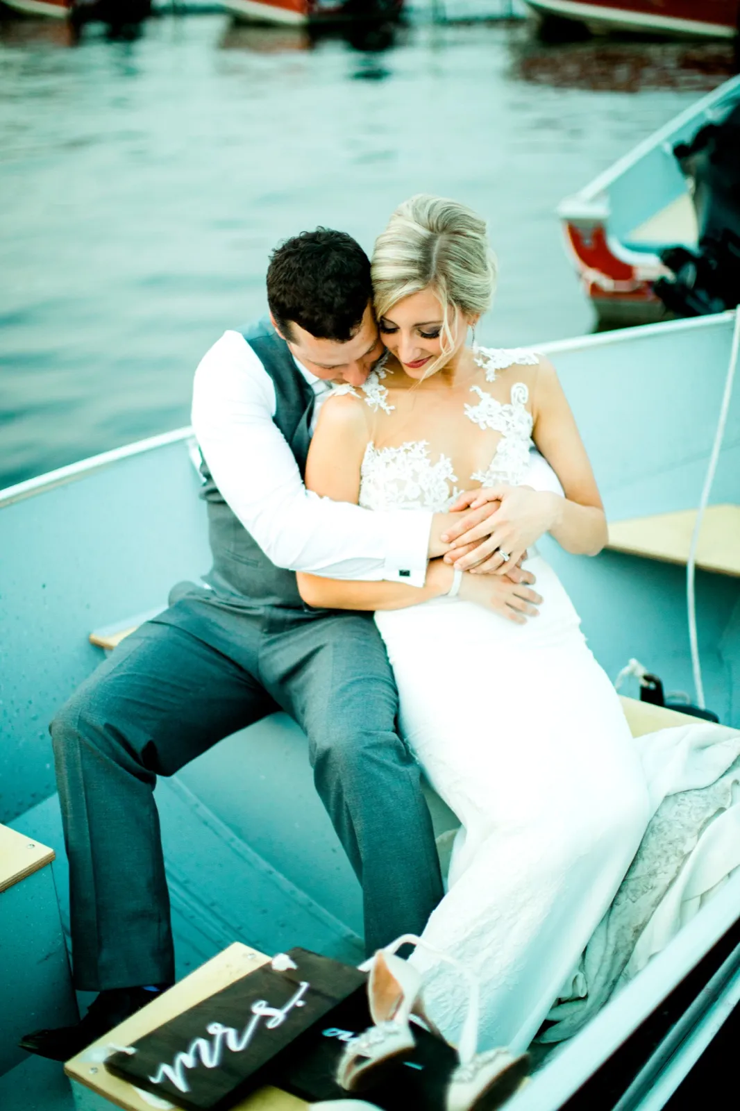 The groom rests his face against the bride's shoulder as they sit together in a small boat on the lake, with a wooden 'mrs' sign and her heeled shoes visible in the foreground. — Tim Larsen Photography, Brainerd Lakes MN