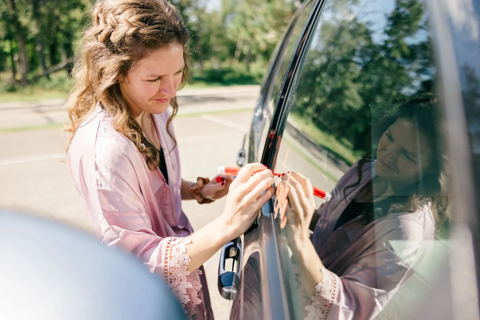 The bride writes a message in marker on a getaway-car window with her reflection visible in the glass — Tim Larsen Photography, Brainerd Lakes MN