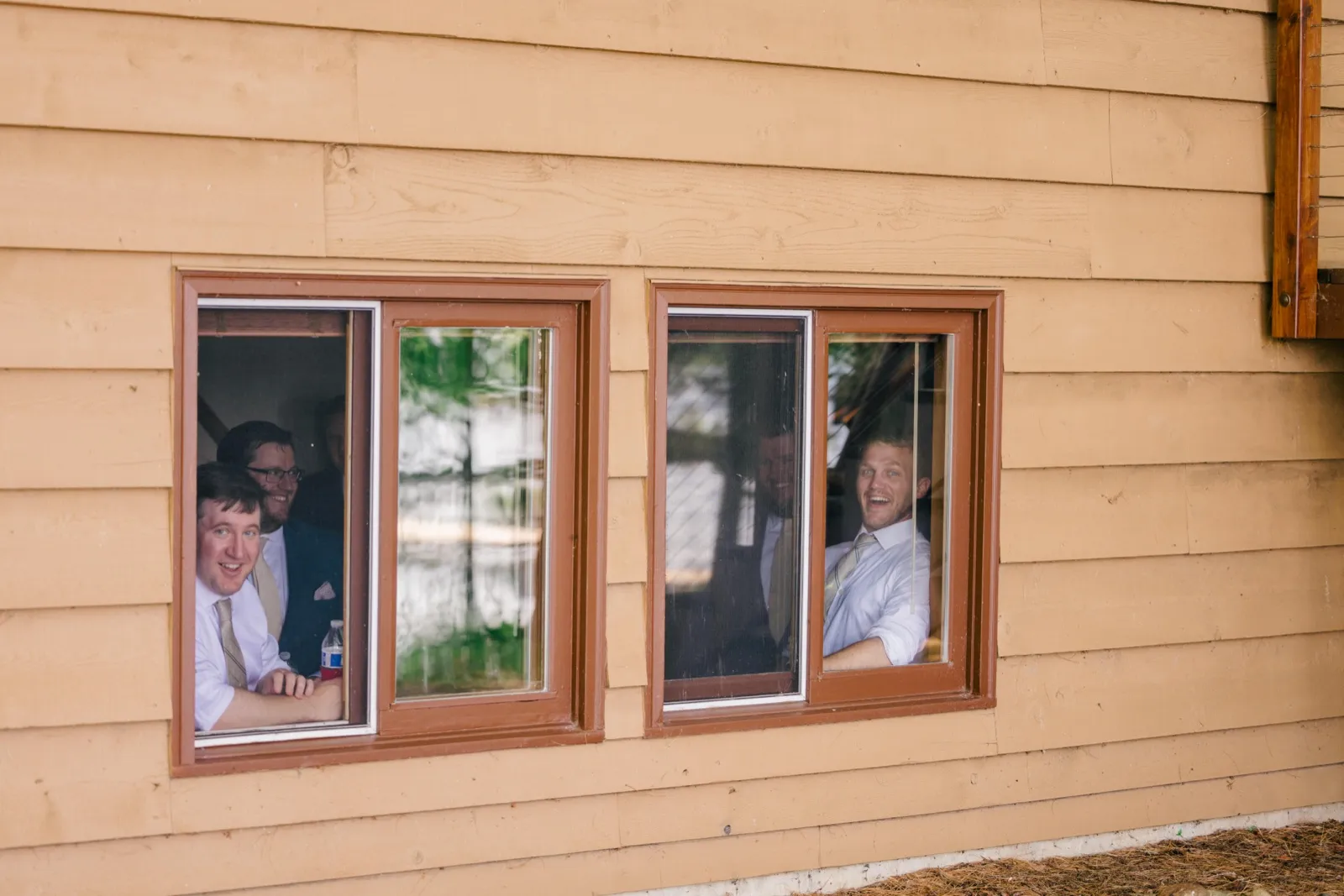 Four groomsmen in shirts and ties peer out through two cabin windows, smiling and laughing at the camera — Tim Larsen Photography, Brainerd Lakes MN
